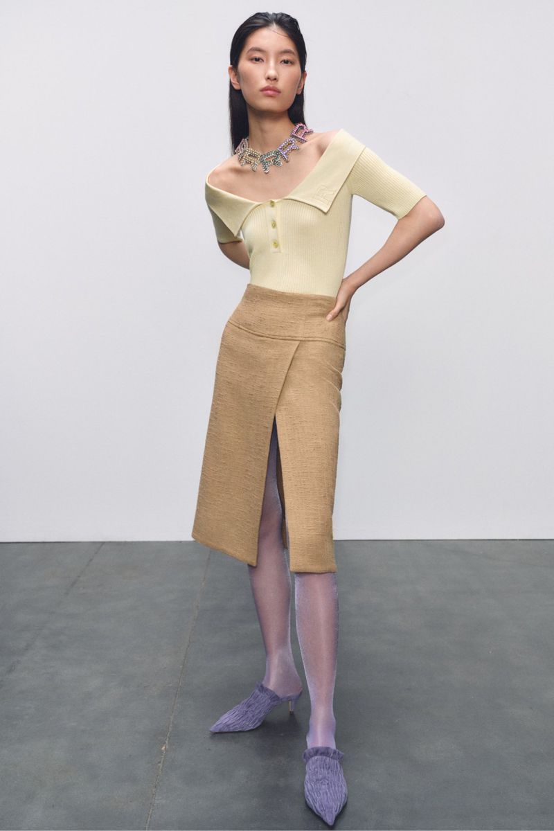 Woman poses in light yellow top and tan midi skirt with a front slit, wearing a statement necklace and lavender heels, against a grey studio backdrop.