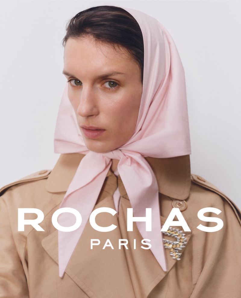 Fashion portrait of a woman with a light pink scarf tied around her head, wearing a trench coat and a jewel brooch.