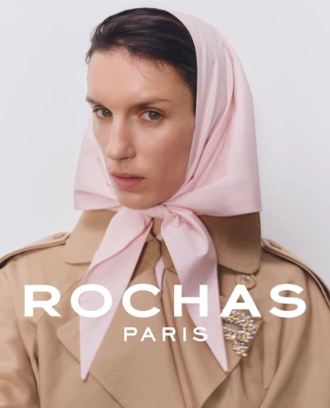 Fashion portrait of a woman with a light pink scarf tied around her head, wearing a trench coat and a jewel brooch.