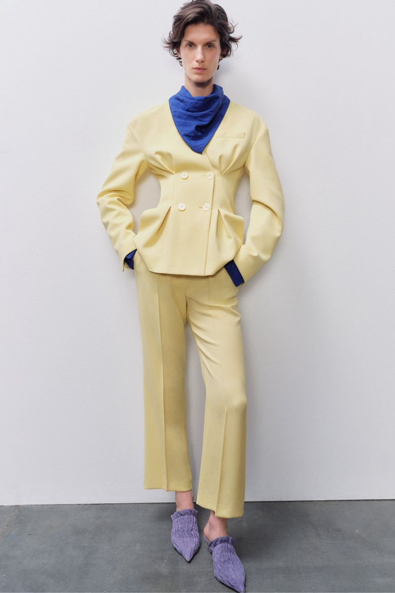 A woman wearing a yellow blazer and pants with blue neckwear and purple shoes poses against a white wall and gray floor in studio lighting.
