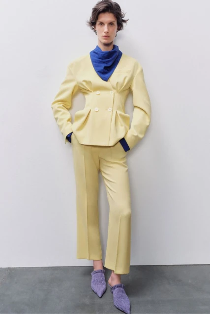 A woman wearing a yellow blazer and pants with blue neckwear and purple shoes poses against a white wall and gray floor in studio lighting.