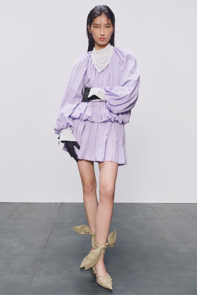 A woman in a lavender dress and strappy heels poses against a neutral background, presenting a high-fashion editorial look in a studio setting.