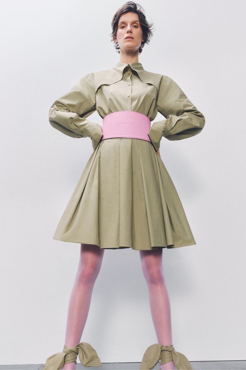 Fashion photo of a woman in a beige dress with a pink belt and matching shoes, posing against a white background in a studio setting.