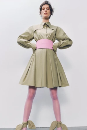 Fashion photo of a woman in a beige dress with a pink belt and matching shoes, posing against a white background in a studio setting.