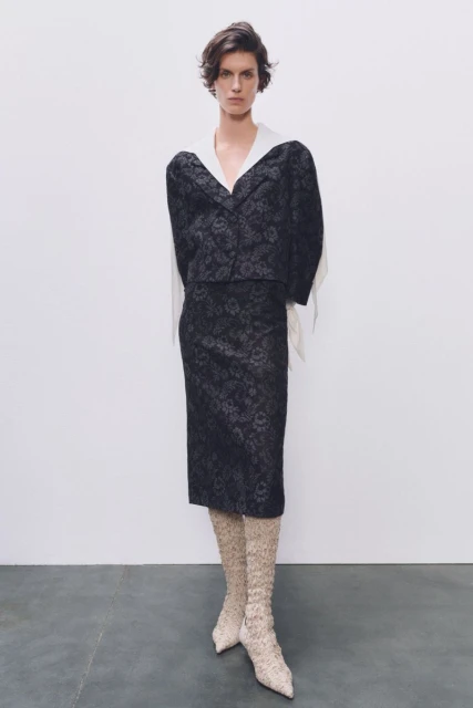 Fashionable woman posing in a studio wearing a floral pattern blazer and skirt with matching floral knee-high boots against a white backdrop.
