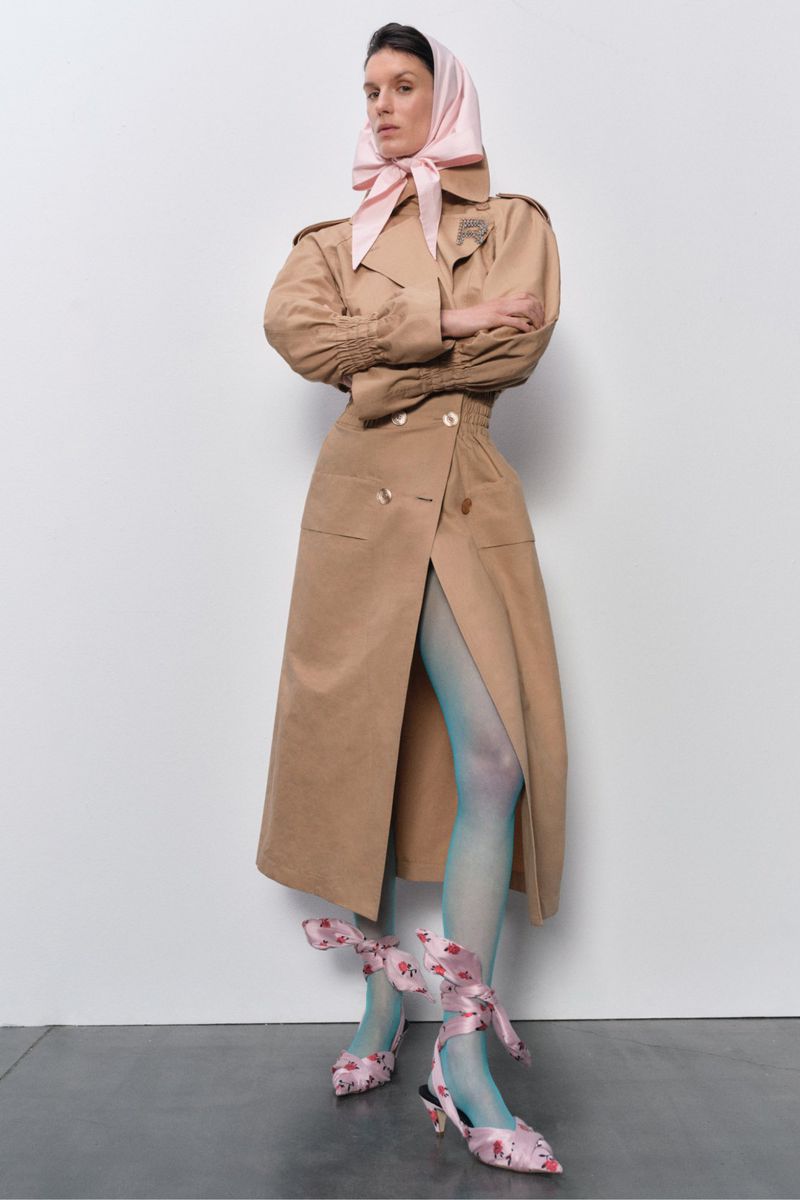 A woman models a tan trench coat, light blue tights, and floral heels with a pink headscarf, posing in front of a white background.