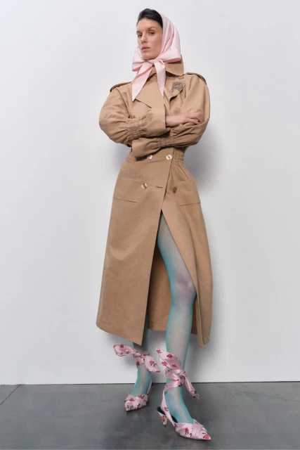 A woman models a tan trench coat, light blue tights, and floral heels with a pink headscarf, posing in front of a white background.