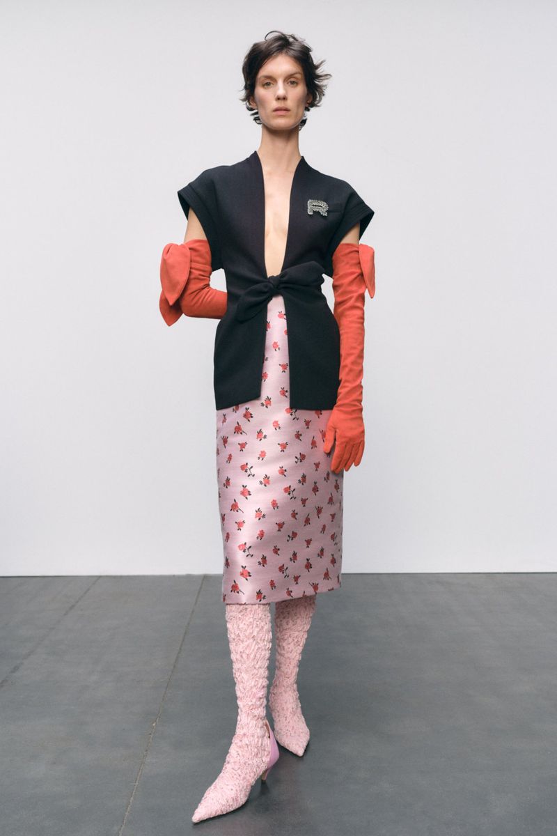 A woman in a black short-sleeved jacket, floral skirt, and pink knit boots stands against a neutral wall, fashion editorial.
