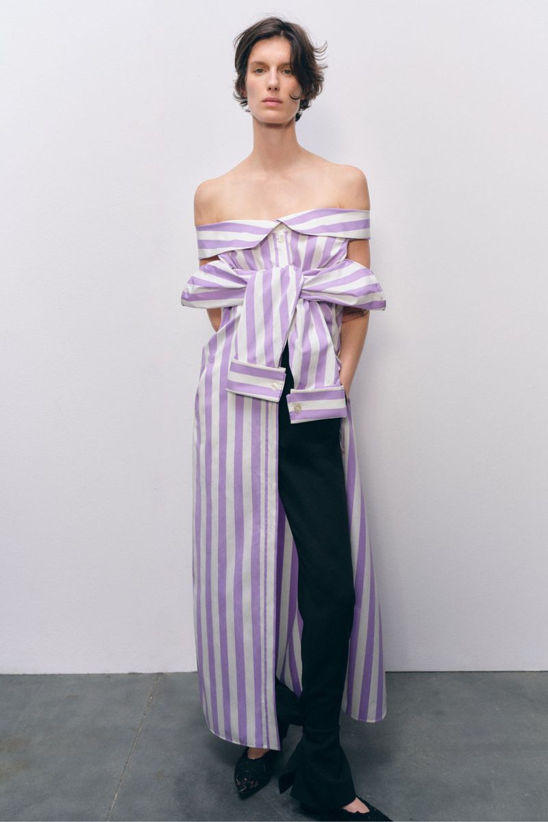 Fashion shot of a woman posing indoors wearing a striped dress over black pants and black pointed-toe shoes against a neutral backdrop.
