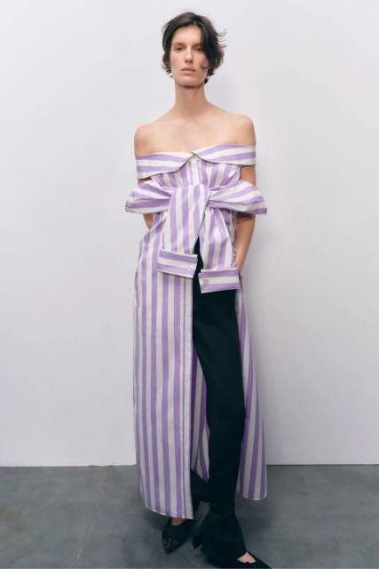 Fashion shot of a woman posing indoors wearing a striped dress over black pants and black pointed-toe shoes against a neutral backdrop.