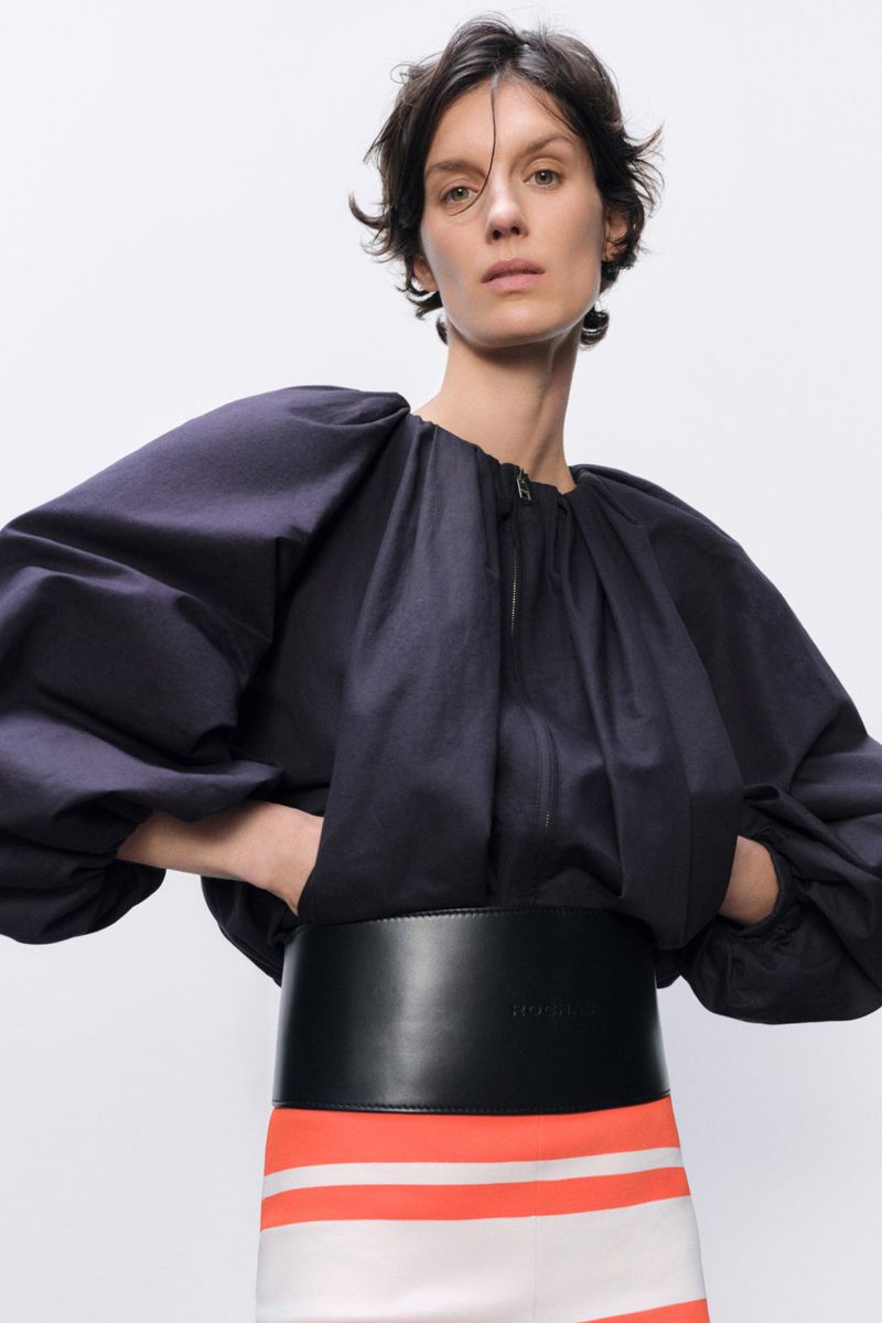 A brunette woman in a fashion pose wears a voluminous-sleeved black blouse, a wide black belt, and a white skirt with orange stripes against a white background.