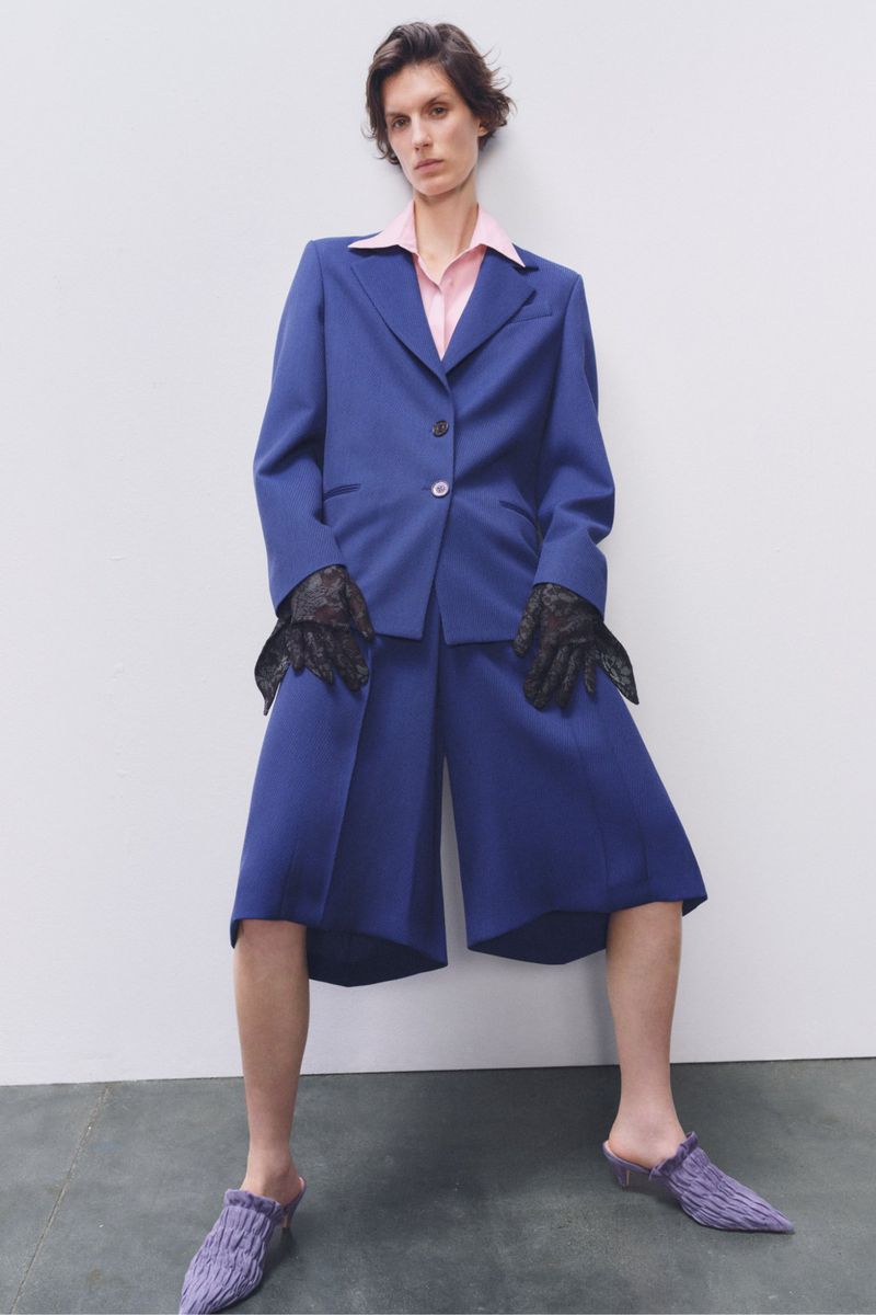 A model poses in a blue suit jacket and shorts with black lace gloves and knit slippers against a white backdrop in a studio shot.