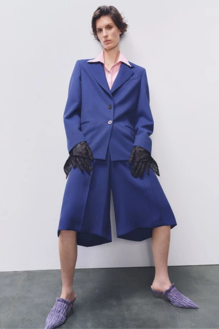A model poses in a blue suit jacket and shorts with black lace gloves and knit slippers against a white backdrop in a studio shot.