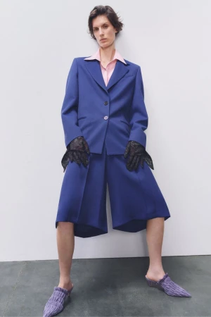 A model poses in a blue suit jacket and shorts with black lace gloves and knit slippers against a white backdrop in a studio shot.