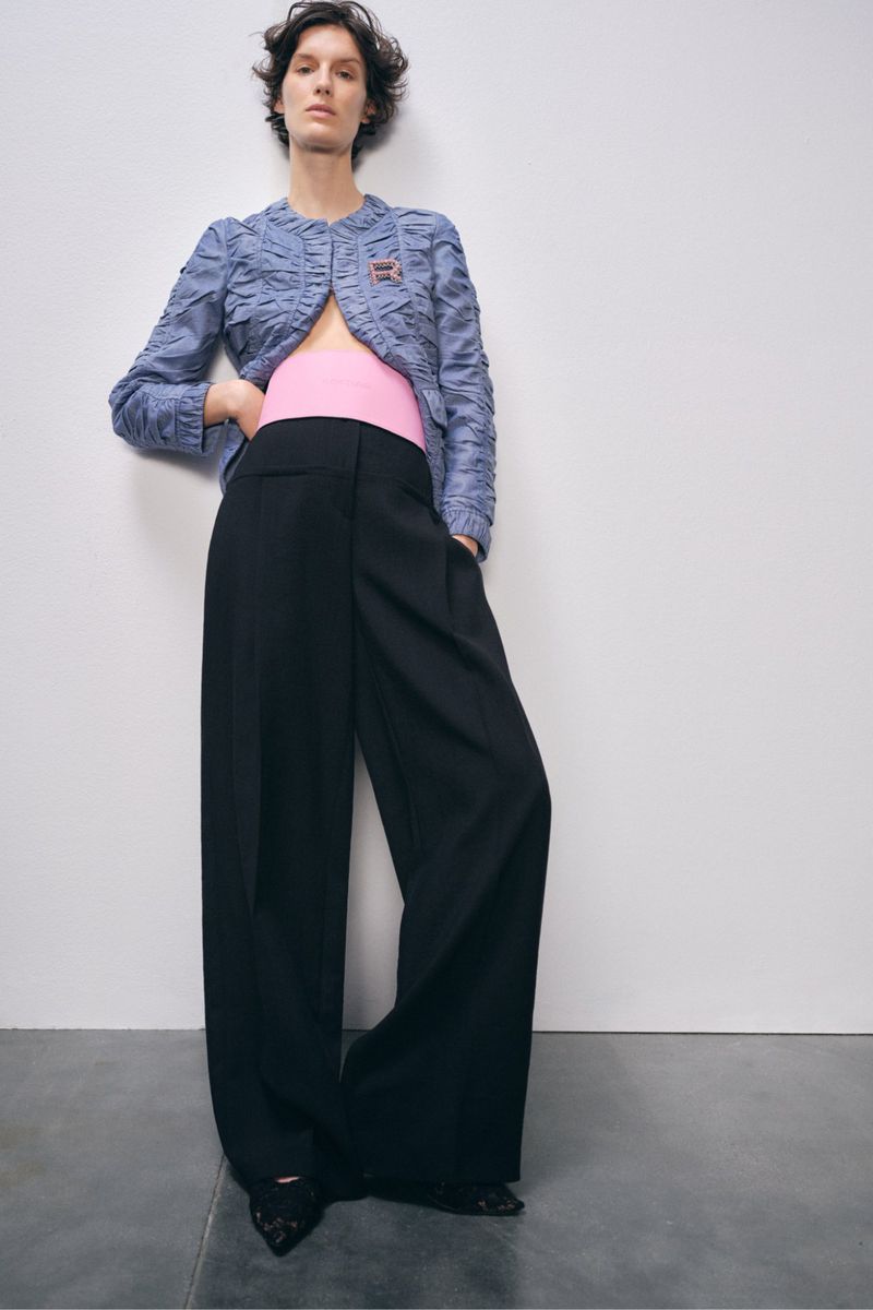 Elegant woman strikes a pose in a blue jacket and black wide-leg trousers against a stark white background, fashion forward.