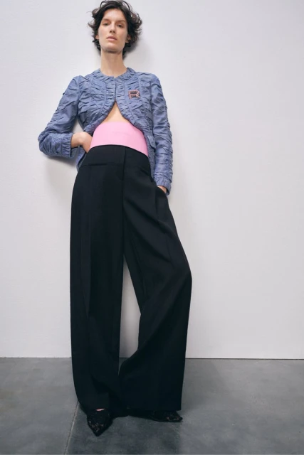 Elegant woman strikes a pose in a blue jacket and black wide-leg trousers against a stark white background, fashion forward.