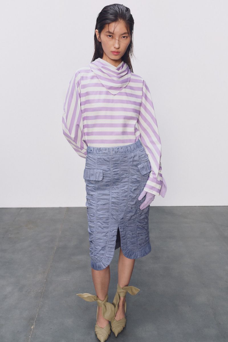 Adult woman poses in a lavender-and-white striped blouse and blue pencil skirt, with ribboned high-heeled shoes, against a white backdrop.