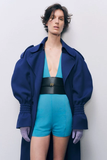 A woman poses in a blue coat and matching shorts with a black belt, complemented by lavender gloves against a neutral background.