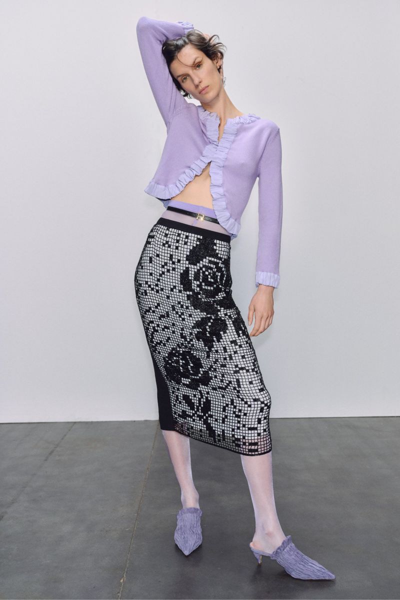 A slender woman poses in a light purple cardigan and black and white rose-patterned skirt against a light grey backdrop in a studio shot.