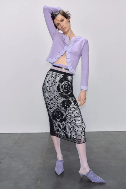 A slender woman poses in a light purple cardigan and black and white rose-patterned skirt against a light grey backdrop in a studio shot.