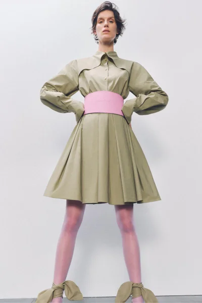 Model in an olive green pleated dress with a wide pink waist belt and matching bow heels against a white backdrop