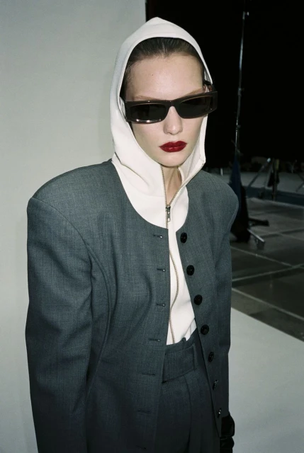 Fashion portrait of a model with bold red lips wearing a grey jacket with a hood and sunglasses against a neutral studio backdrop.