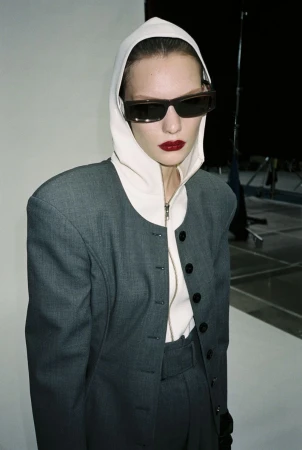 Fashion portrait of a model with bold red lips wearing a grey jacket with a hood and sunglasses against a neutral studio backdrop.