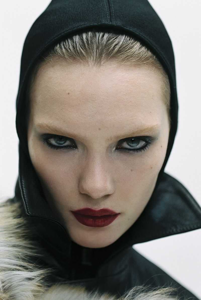 Close-up portrait of a woman with dark eye makeup and dark red lipstick wearing a black hood and fur-trimmed jacket against a white background.