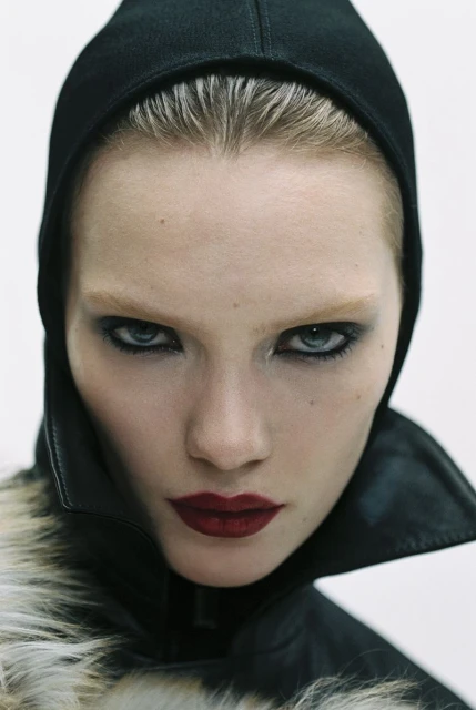 Close-up portrait of a woman with dark eye makeup and dark red lipstick wearing a black hood and fur-trimmed jacket against a white background.