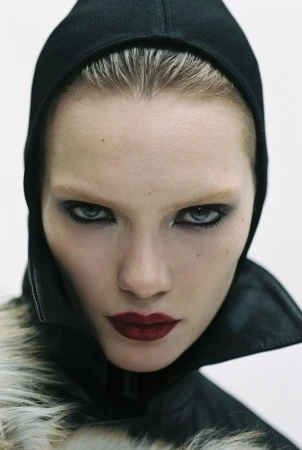 Close-up portrait of a woman with dark eye makeup and dark red lipstick wearing a black hood and fur-trimmed jacket against a white background.
