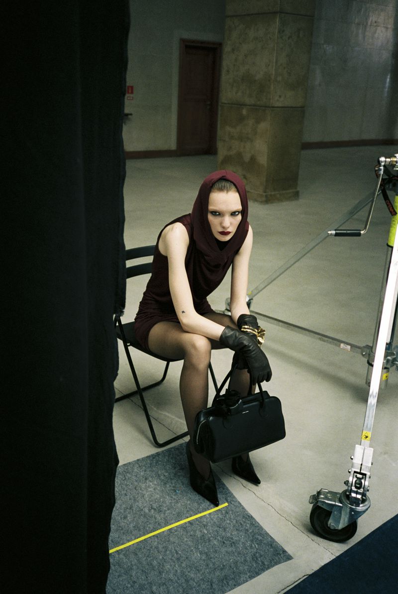 Model in a burgundy hooded sleeveless mini dress with black gloves and handbag seated on a folding chair in a studio.