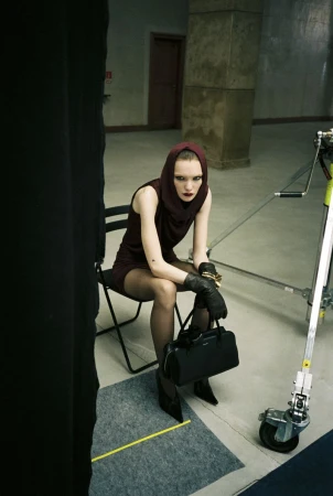 Model in a burgundy hooded sleeveless mini dress with black gloves and handbag seated on a folding chair in a studio.
