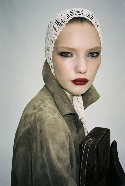Close-up fashion shot of a woman wearing a crochet bonnet, suede coat, leather gloves, and holding a brown handbag in a studio setting.