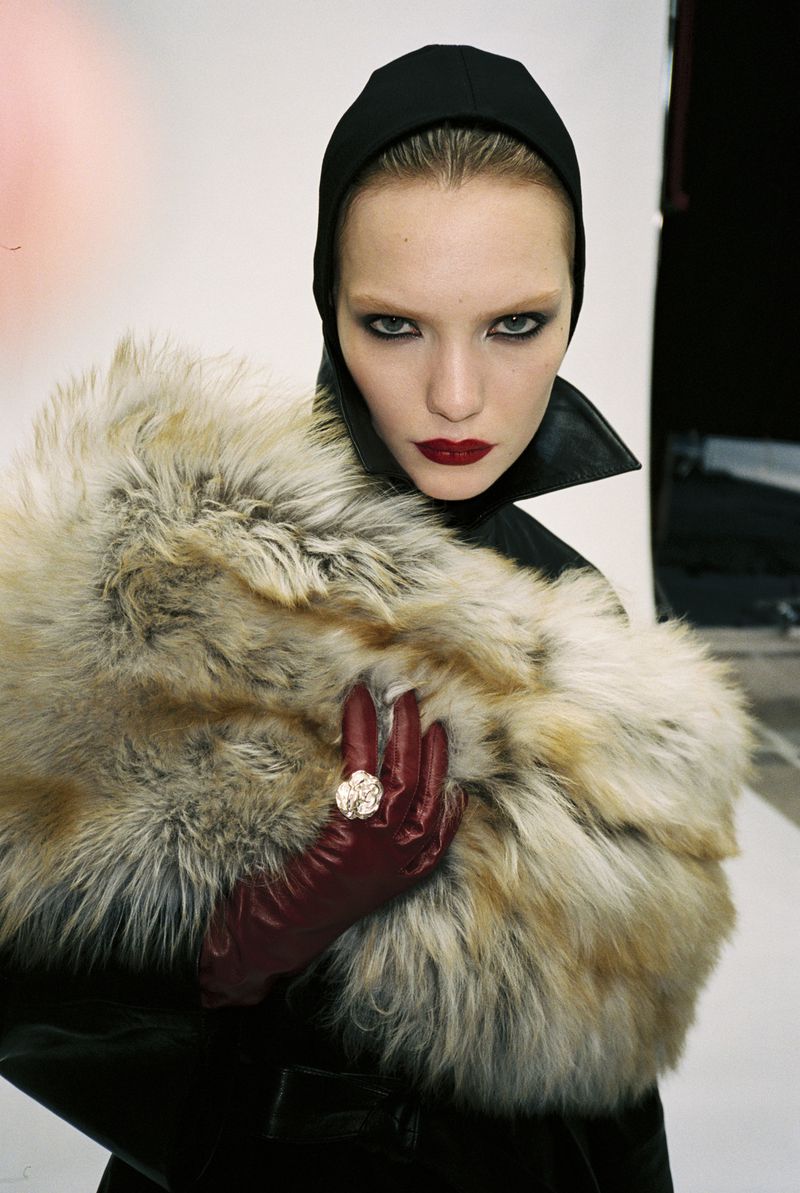 Fashion portrait of a woman with bold makeup wearing a black hood, leather coat, fur stole, and burgundy gloves with a large ring indoors.