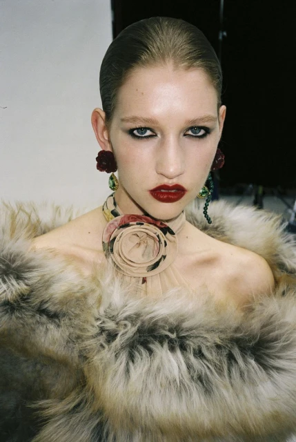 Elegant portrait of an adult female fashion model with dark red lipstick, wearing a fur coat and a floral necklace and earrings.