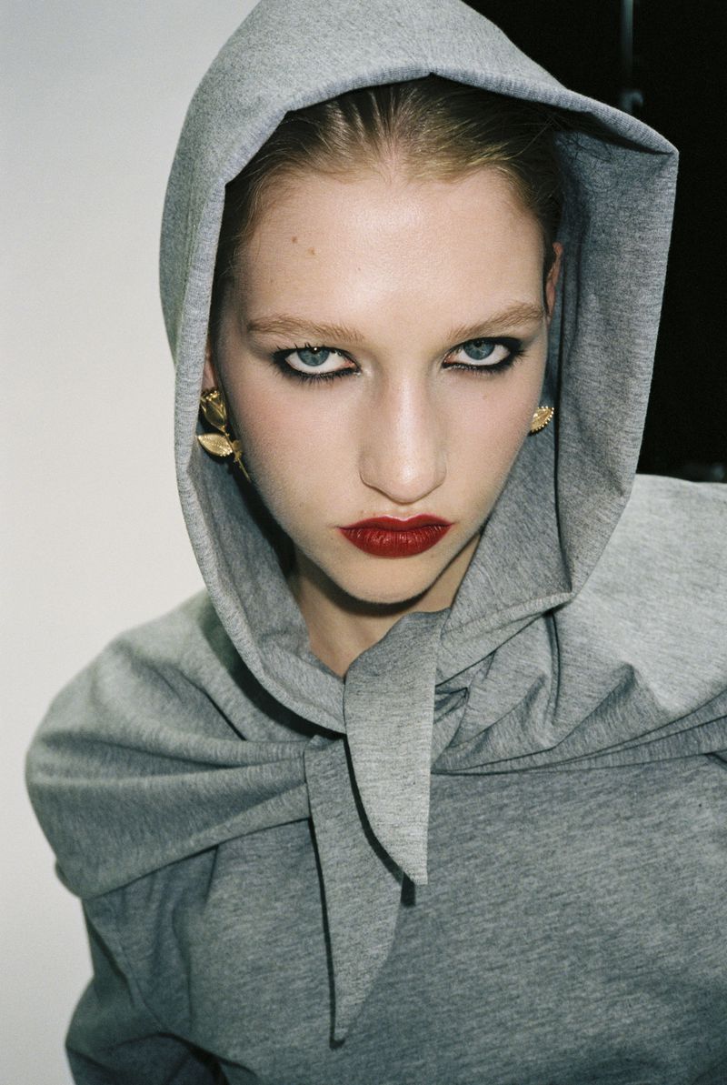 Close-up of a woman with red lipstick wearing a grey hooded top, her intense gaze and golden earrings adding to the portrait's allure.