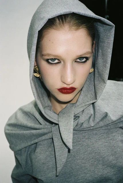 Close-up of a woman with red lipstick wearing a grey hooded top, her intense gaze and golden earrings adding to the portrait's allure.