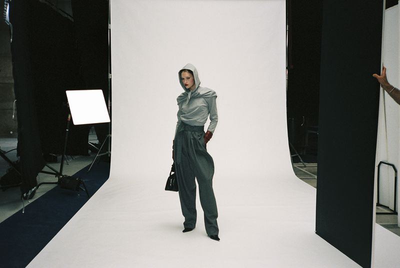 Fashion shoot in photo studio features a woman in grey, hooded top and pleated pants, holding a black purse, under studio lights.