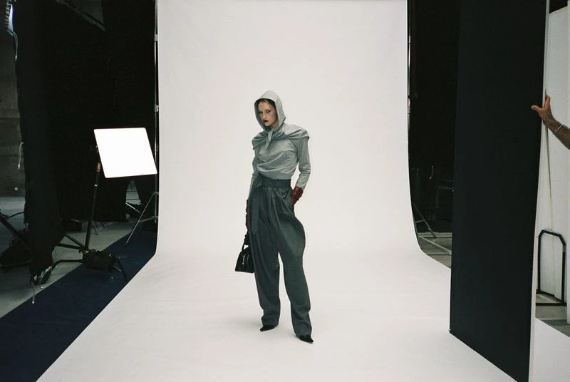 Fashion shoot in photo studio features a woman in grey, hooded top and pleated pants, holding a black purse, under studio lights.