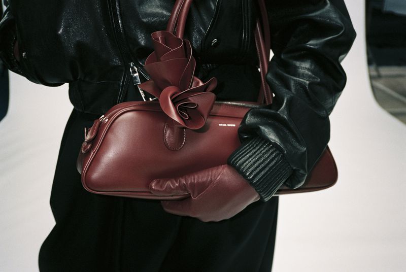 Close-up of a red leather handbag decorated with a red leather flower, held by a person wearing black leather gloves and jacket.
