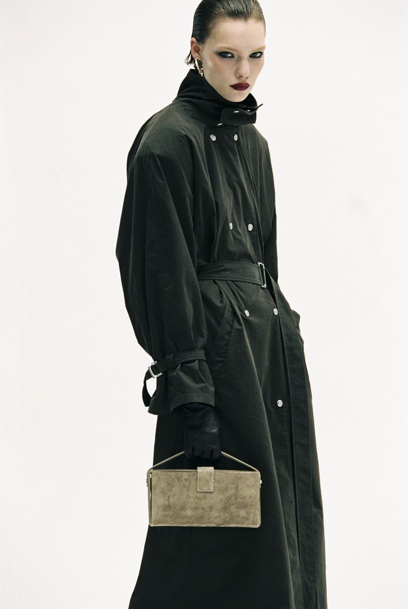 Fashion shot of a woman in a trench coat holding a handbag against a white background in a studio. The shot highlights the texture of the coat.