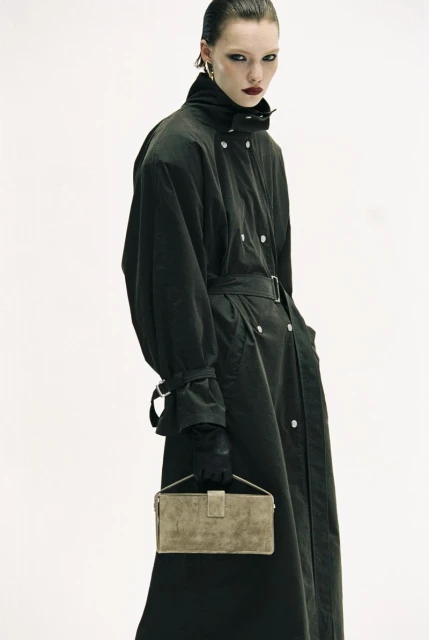 Fashion shot of a woman in a trench coat holding a handbag against a white background in a studio. The shot highlights the texture of the coat.