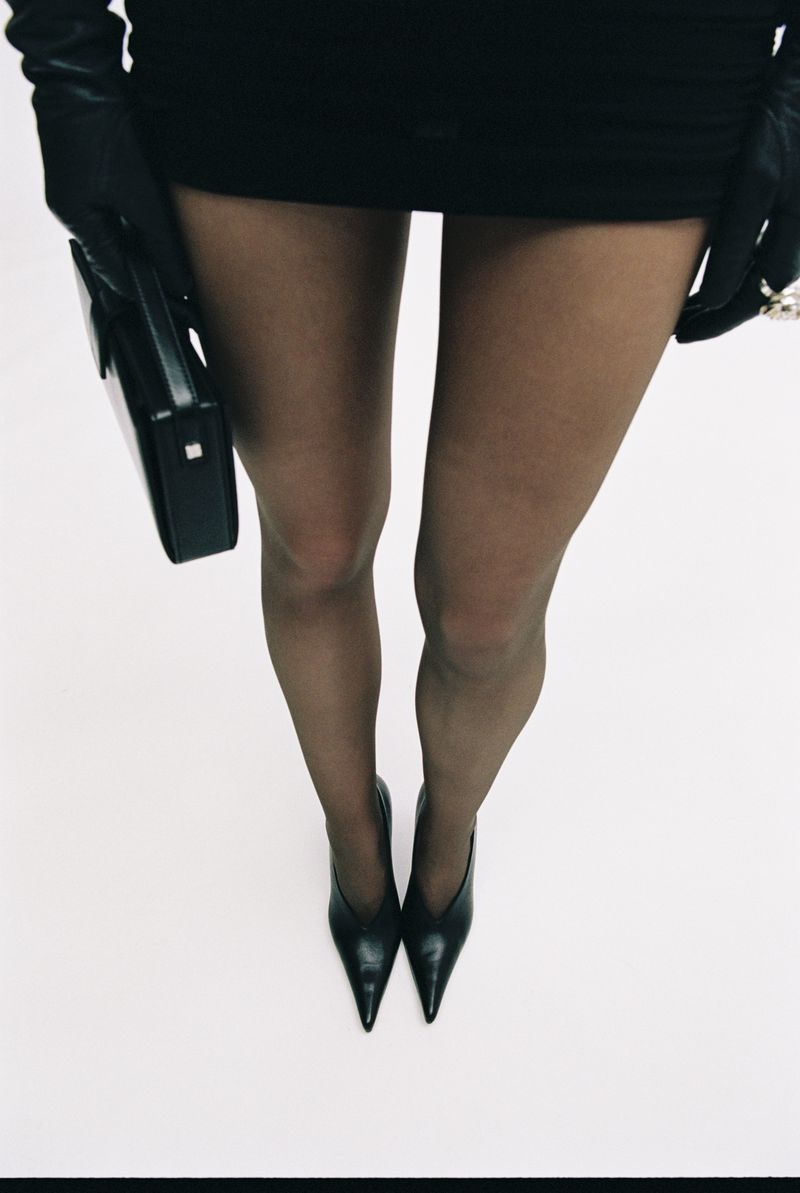 Fashion shot featuring a woman's legs in black tights and pointed-toe heels against a white background, with a black purse and gloves.