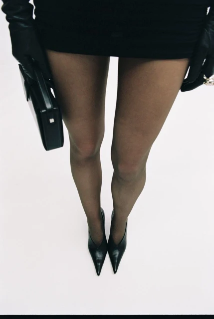 Fashion shot featuring a woman's legs in black tights and pointed-toe heels against a white background, with a black purse and gloves.