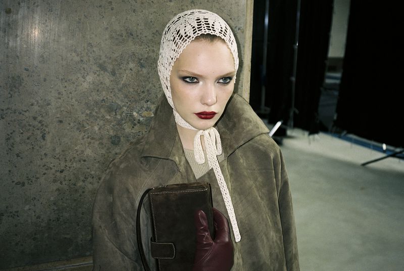 Woman in a vintage bonnet and coat poses indoors with a purse. The neutral color palette enhances the fashion editorial style.