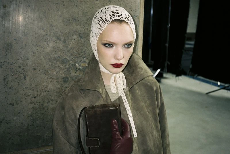 Woman in a vintage bonnet and coat poses indoors with a purse. The neutral color palette enhances the fashion editorial style.