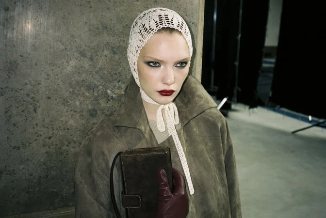 Woman in a vintage bonnet and coat poses indoors with a purse. The neutral color palette enhances the fashion editorial style.