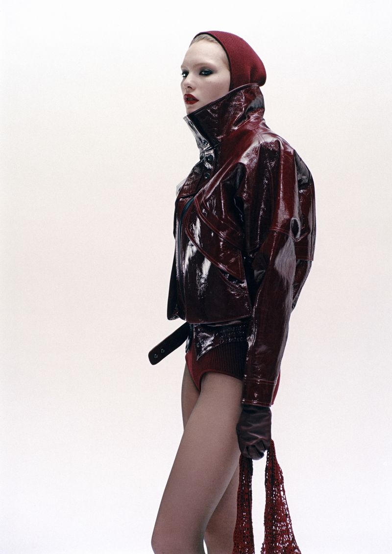 A model poses in a shiny red vinyl jacket with a hood, long gloves, and a matching scarf against a white background in a studio setting.