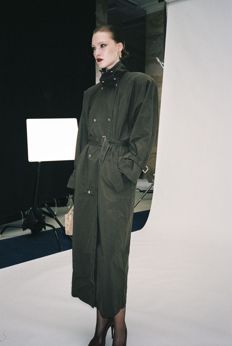 A fashionably dressed woman in a long green trench coat poses in a photo studio with a white backdrop and studio lighting for a portrait.