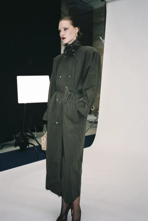 Model wearing a long dark khaki belted trench coat with a high collar and pointed heels against a studio backdrop.